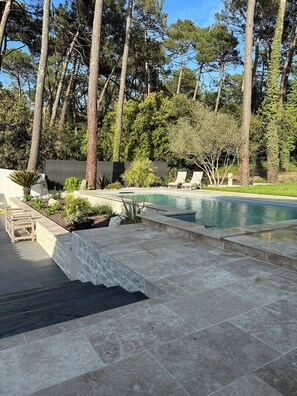 Une piscine dotée d’une plage en pierre, entourée de grands arbres et bordée d’une pelouse.