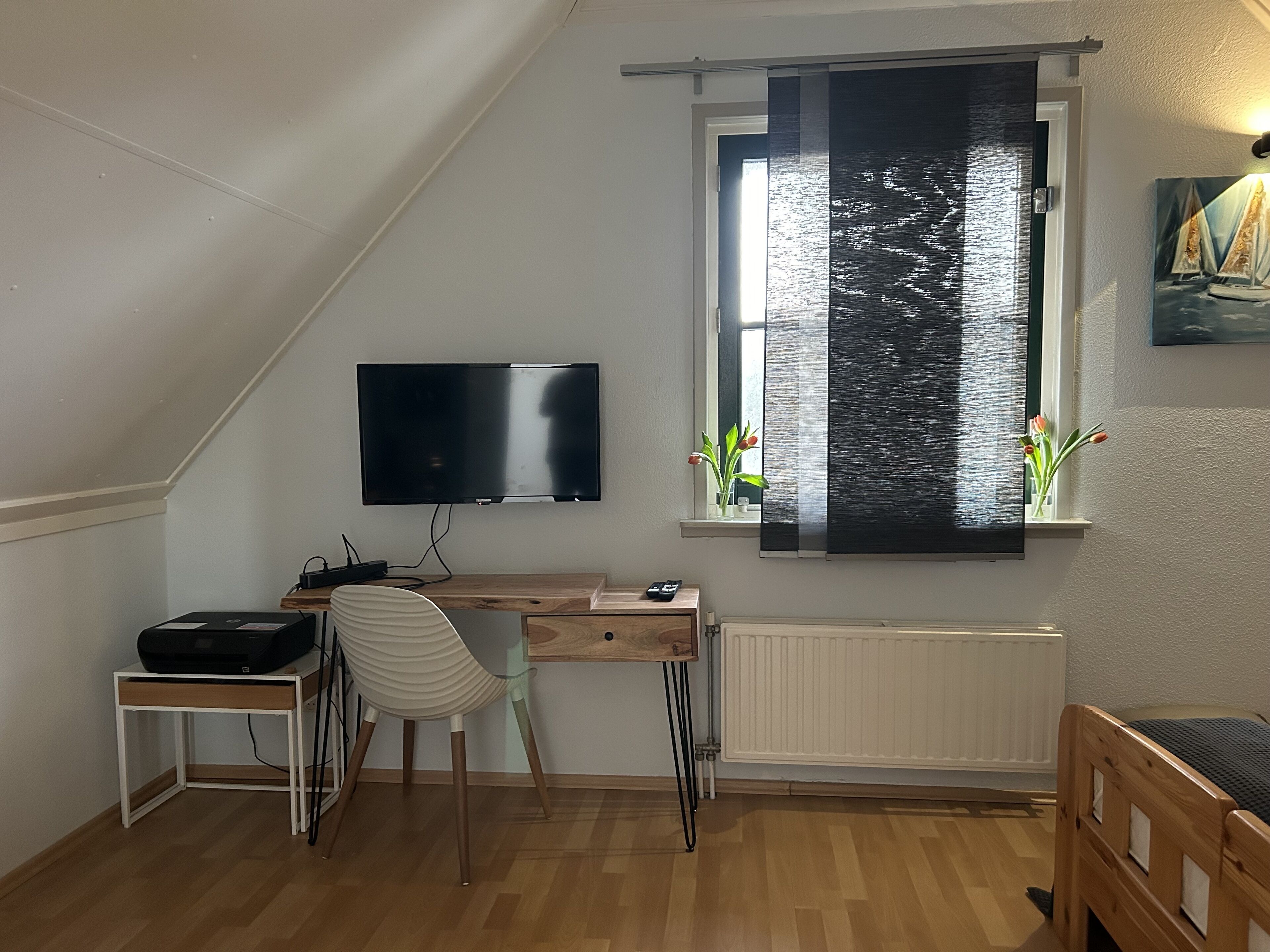 Une pièce avec un bureau en bois, une chaise, un téléviseur fixé au mur, un radiateur et une fenêtre donnant sur l’eau.