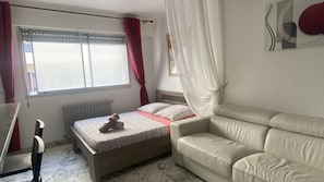A bedroom with a bed, a beige sofa, a radiator, a window with curtains, and a wall with a picture.