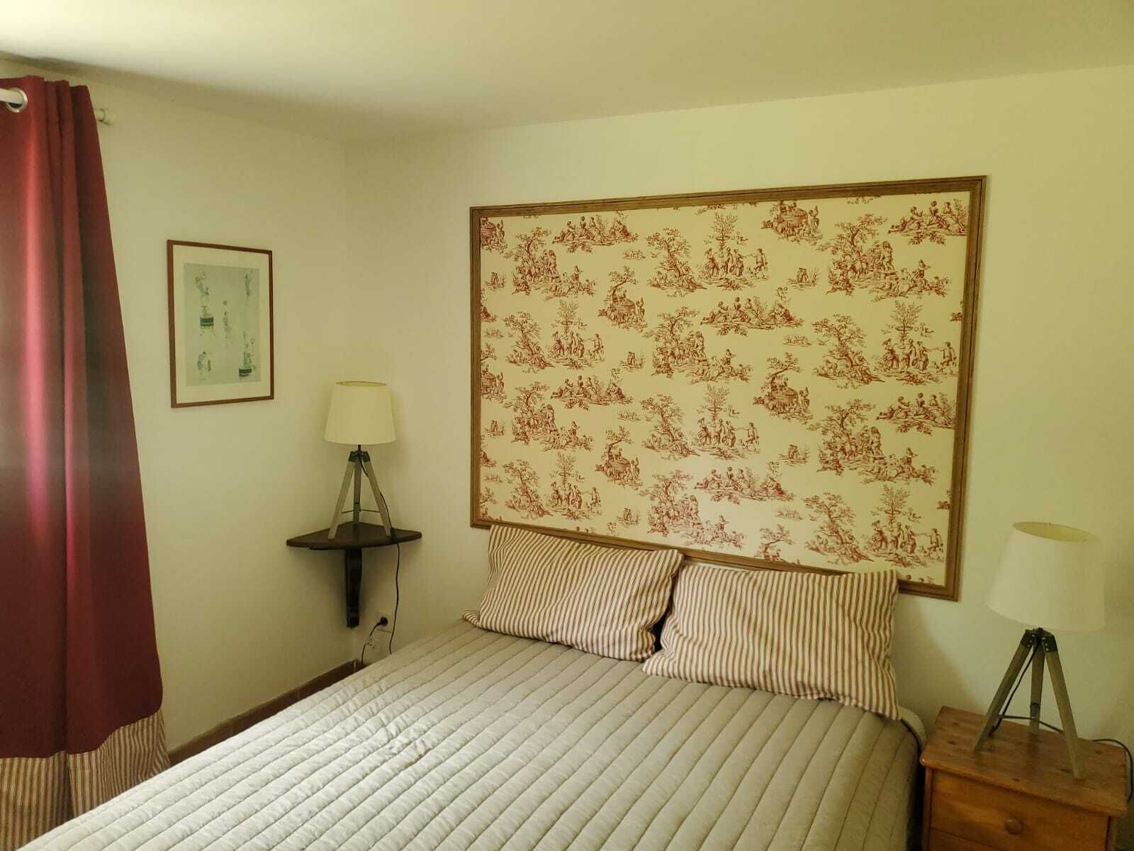 A bedroom with a bed, striped pillows, a nightstand with a lamp, a framed picture, and a red curtain.