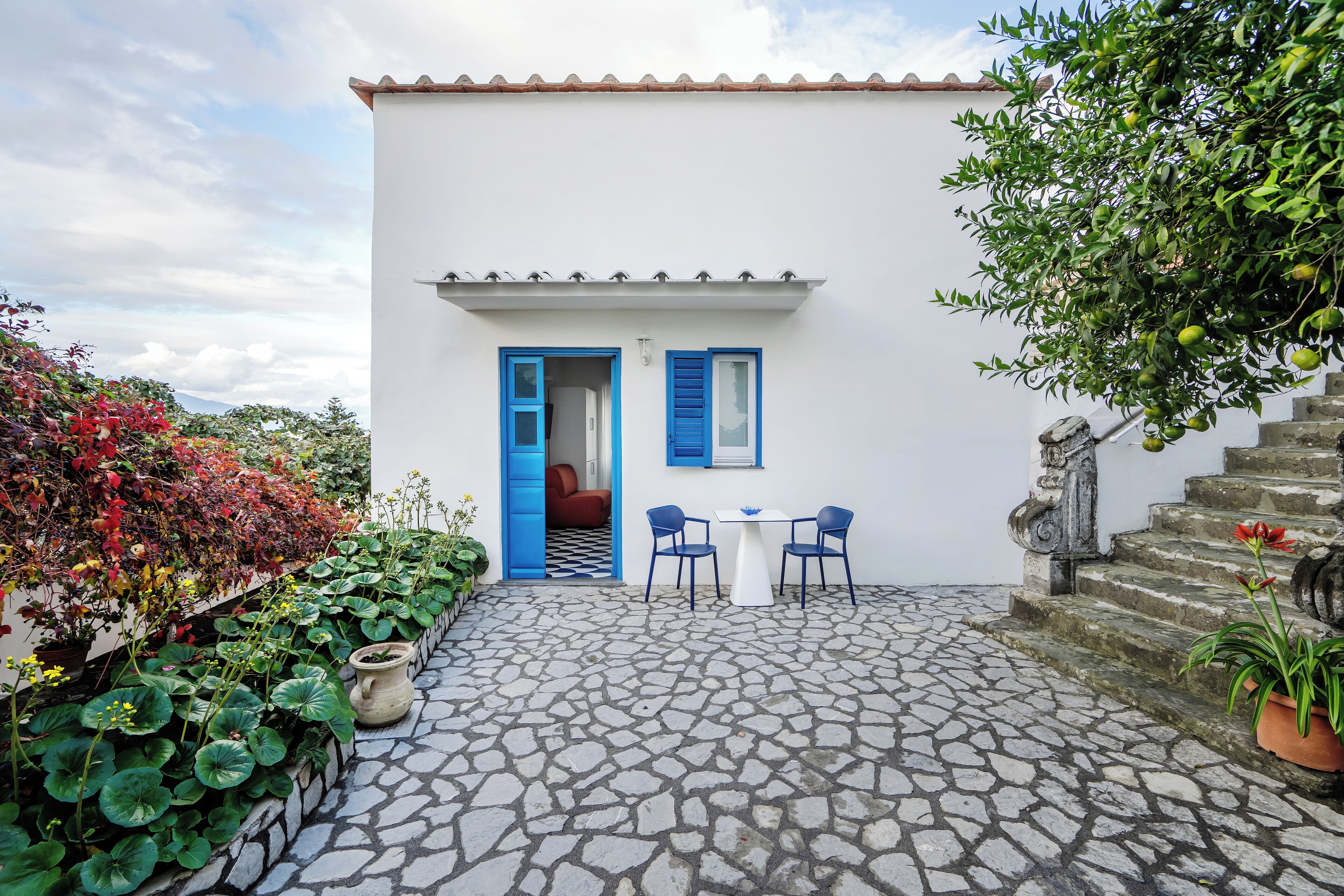 A white building with blue shutters, a stone-paved courtyard, and a small table with chairs.