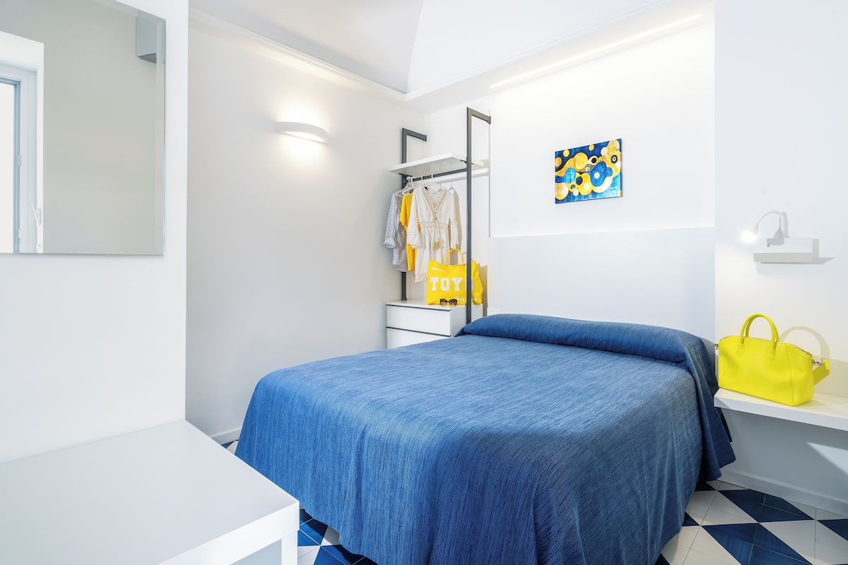 A modern bedroom with a bed, a nightstand, a mirror, a hanging rack with clothes, a yellow bag, and a painting on the wall.