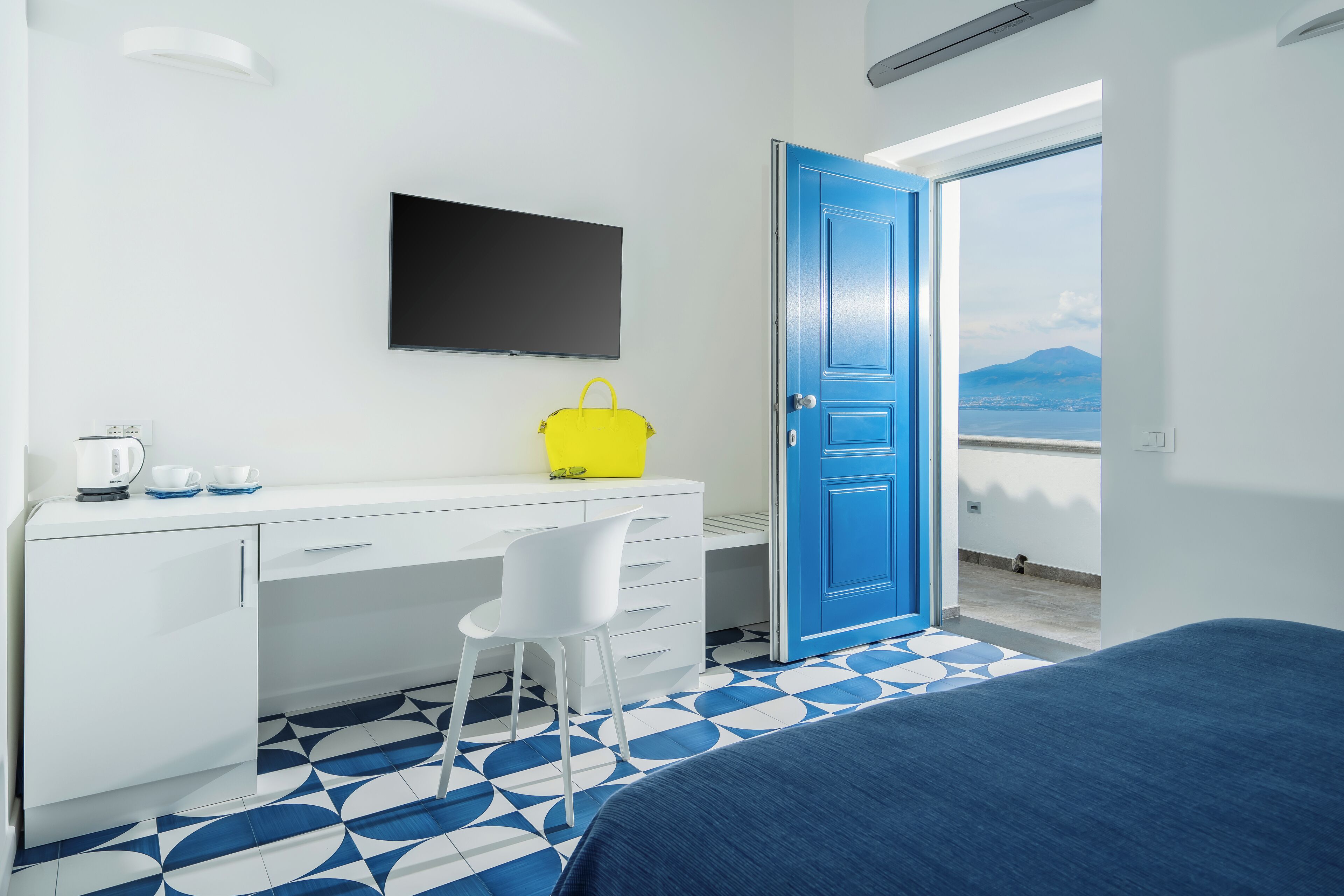 A hotel room with a bed, a desk with a chair, a TV, a bright yellow handbag, and a blue door leading to a balcony with a view of a mountain.