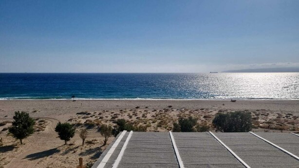 Una playa con un área pavimentada en primer plano, vista despejada del océano y un barco distante en el horizonte.
