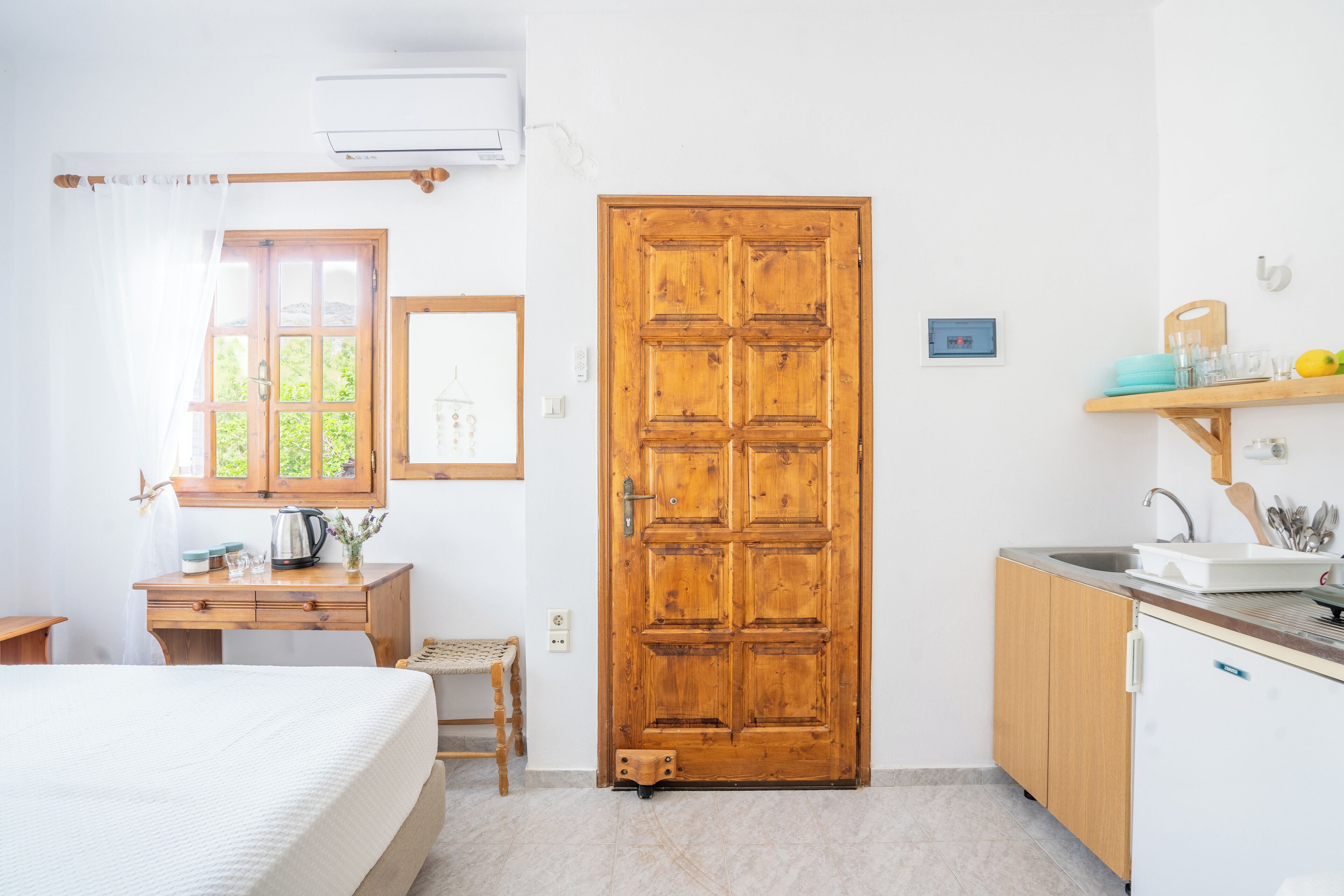 Una habitación con puerta de madera, cama, escritorio, mesita, estante con utensilios de cocina y ventana con cortinas.