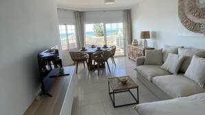 Ein modernes Wohnzimmer mit Sofa, Couchtisch und Esstisch. Durch große Fenster hat man Blick auf den Ozean.