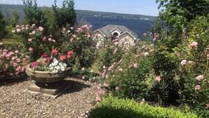 A garden with pink roses and a stone planter, a house with a grey roof, and a view of the water and hills in the background.
