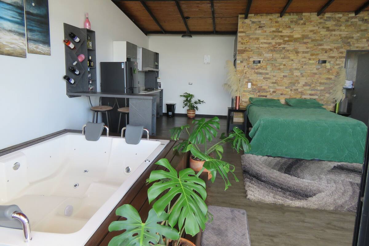 A modern room with a large bathtub, a bed with a green blanket, and a stone wall.