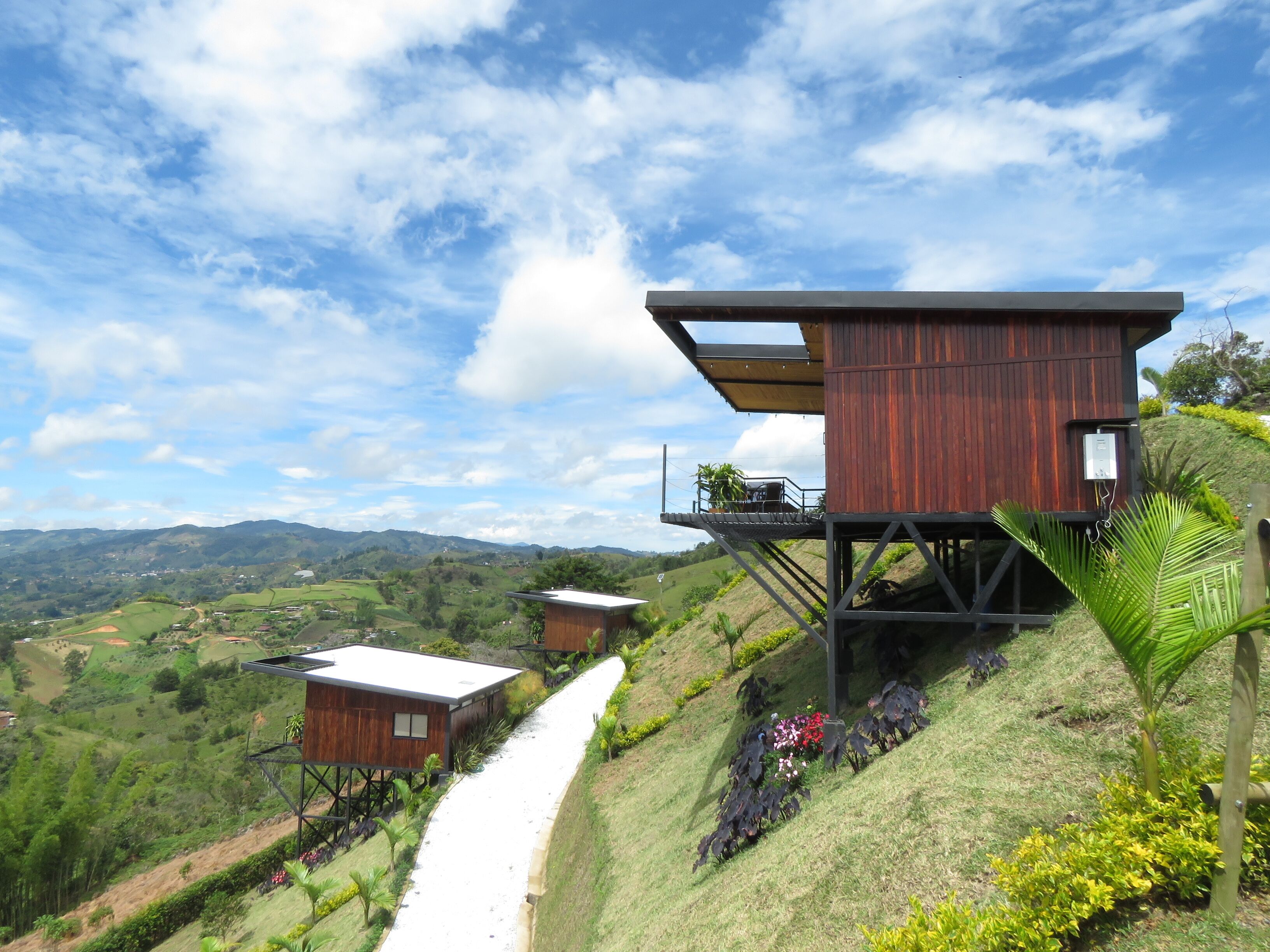A modern wooden cabin with a balcony, situated on a hillside with a pathway leading to it.
