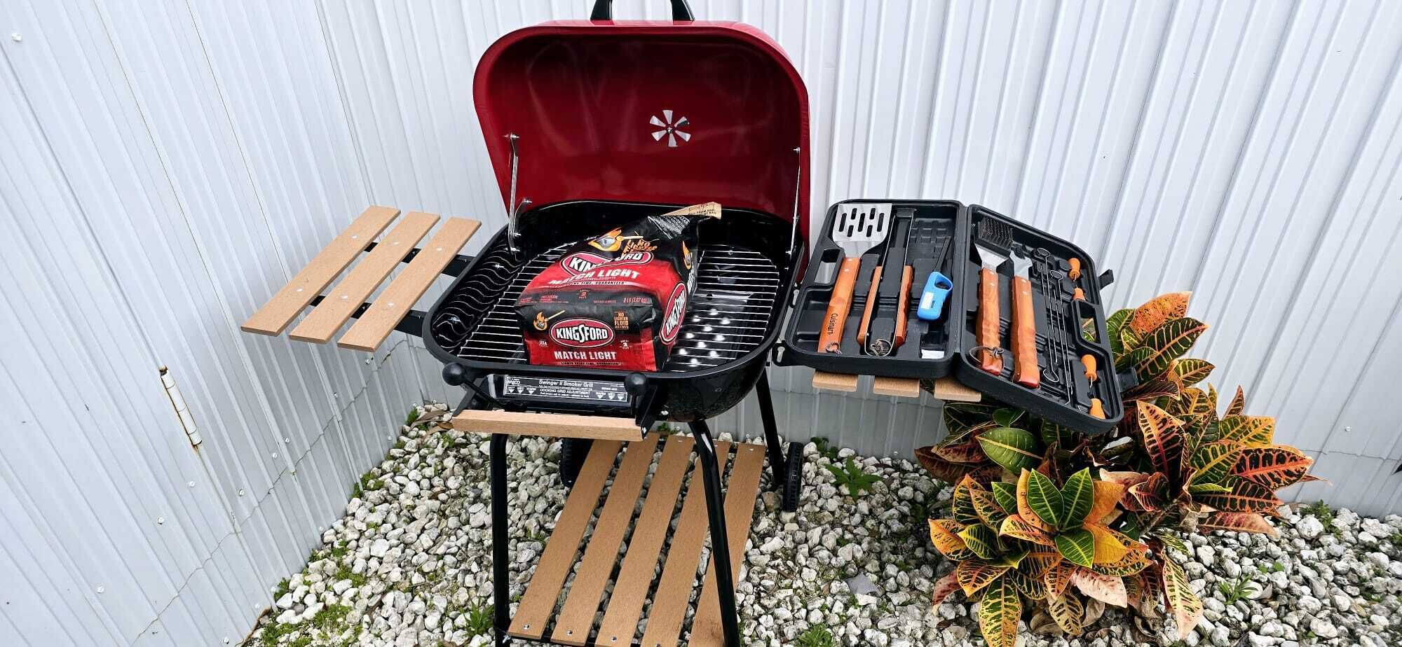 Een draagbare grill met een rode hoes, een zak houtskool en een set grillgereedschap.