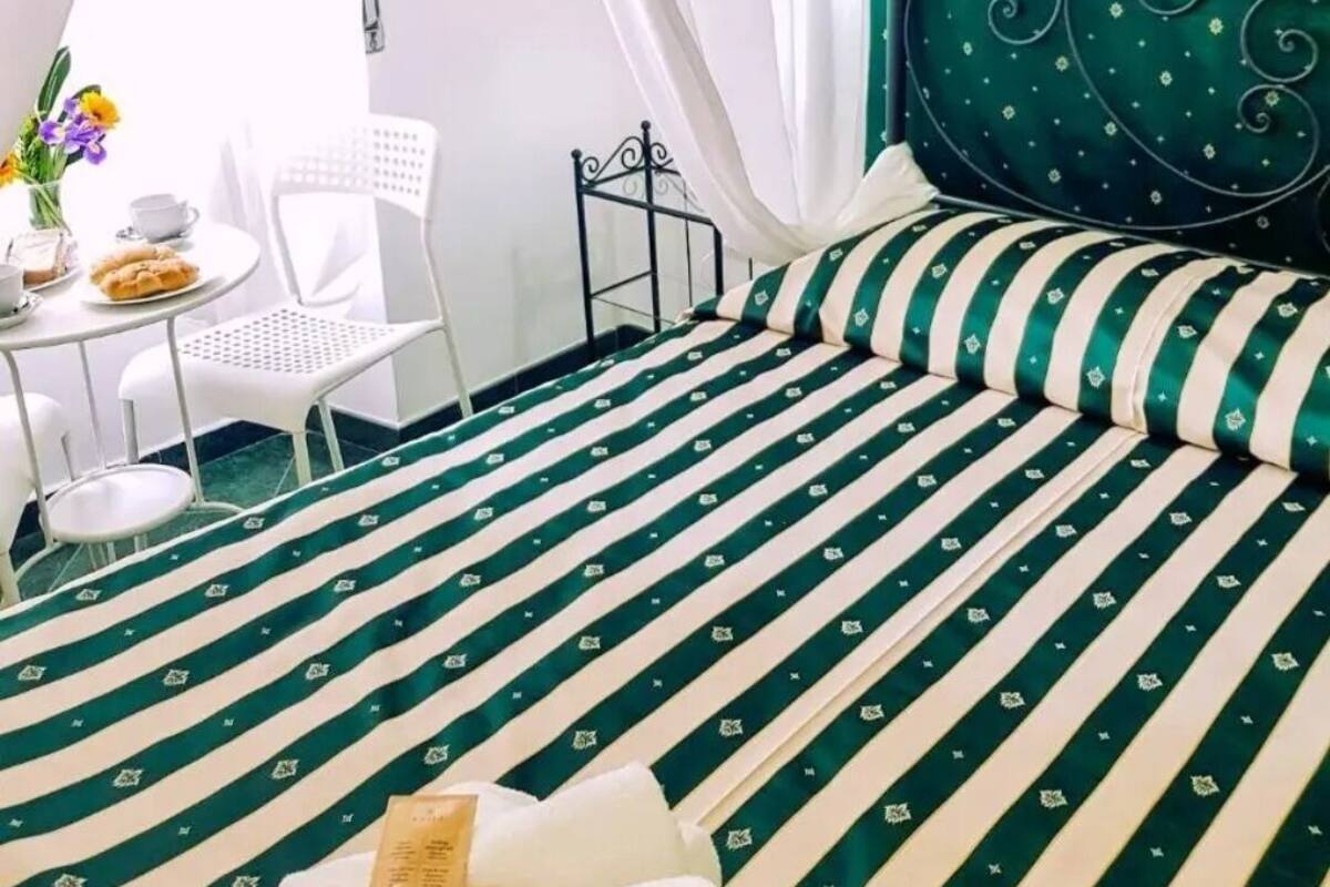 Een net opgemaakt bed met gestreepte lakens, een tafeltje met een bloevas en een stoel.