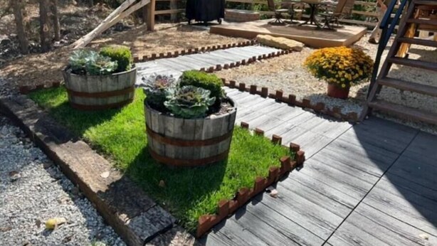 Una terraza de madera con zona de barbacoa, mobiliario exterior y jardineras con plantas.