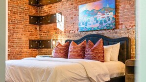A bed with a dark headboard, white bedding, and patterned pillows, set against a brick wall with a painting above.