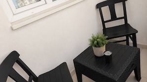 A small outdoor seating area with two black chairs and a square table, a potted plant, and a window with white trim. - Brand otel (Didim)