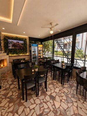 A restaurant with stone-tiled flooring, black tables and chairs, a ceiling fan, a television mounted on a wall, and a Pepsi refrigerator. - Brand otel (Didim)