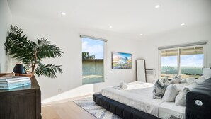 Une chambre moderne avec un grand lit, un canapé foncé, une grande fenêtre donnant sur la ville, une plante en pot et un tableau accroché au mur.