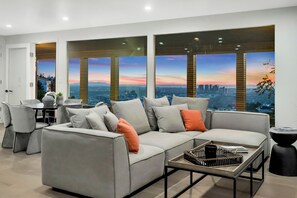 Un moderno salón con un sofá gris, una mesa de centro y vistas a un paisaje urbano a través de amplios ventanales.