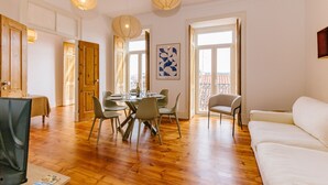 A spacious living area with a dining table set for four, surrounded by chairs. There are two large windows with a view of the outside, and a balcony visible through one window. - RENT4REST CASTLE VIEW FAMILY & FRIENDS BAIRRO ALTO (Lisboa ciudad)