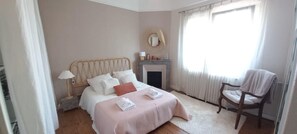 Um quarto com cama, uma cadeira, lareira e janela com cortinas.