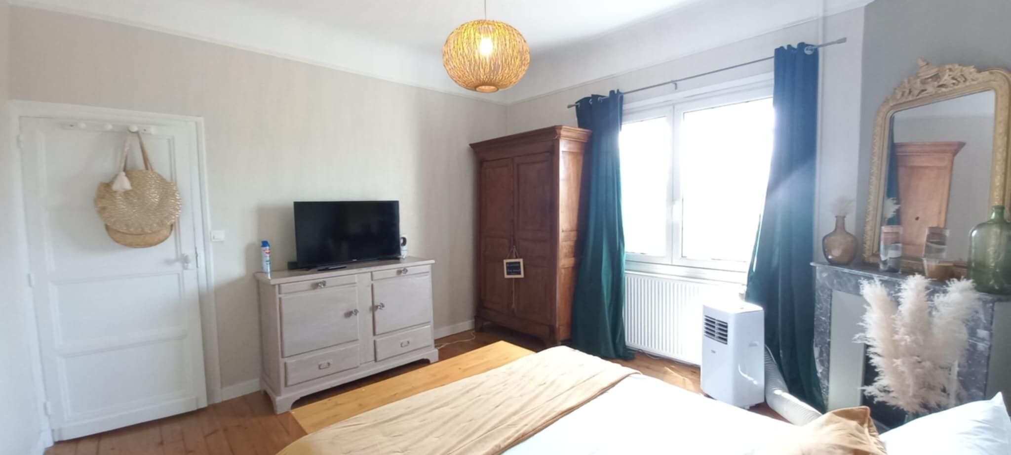 Une chambre à coucher avec un lit, une commode, un téléviseur posé sur la commode, une fenêtre avec des rideaux, un radiateur et un miroir avec un vase.