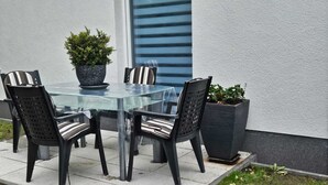 Una mesa con cubierta de vidrio, cuatro sillas, dos plantas en macetas y una ventana con persianas horizontales.
