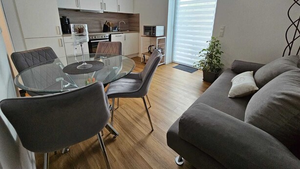 Una sala moderna con una mesa de comedor de vidrio, un sofá gris y una cocina al fondo.
