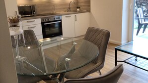 Una cocina moderna con horno integrado, microondas y electrodomésticos de acero inoxidable. Hay una mesa de vidrio para comer con dos sillas y una pequeña planta en maceta sobre la encimera.