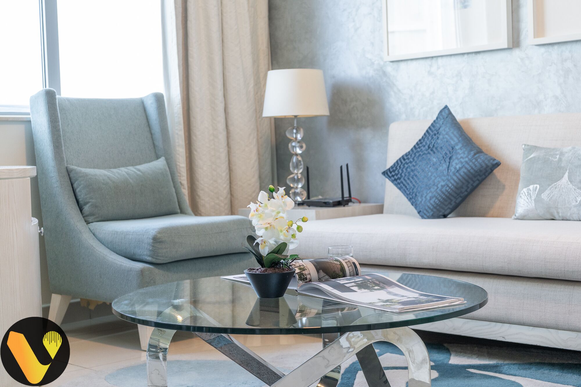 Een moderne woonkamer met een glazen salontafel, een bank met een blauw kussen en een lamp.