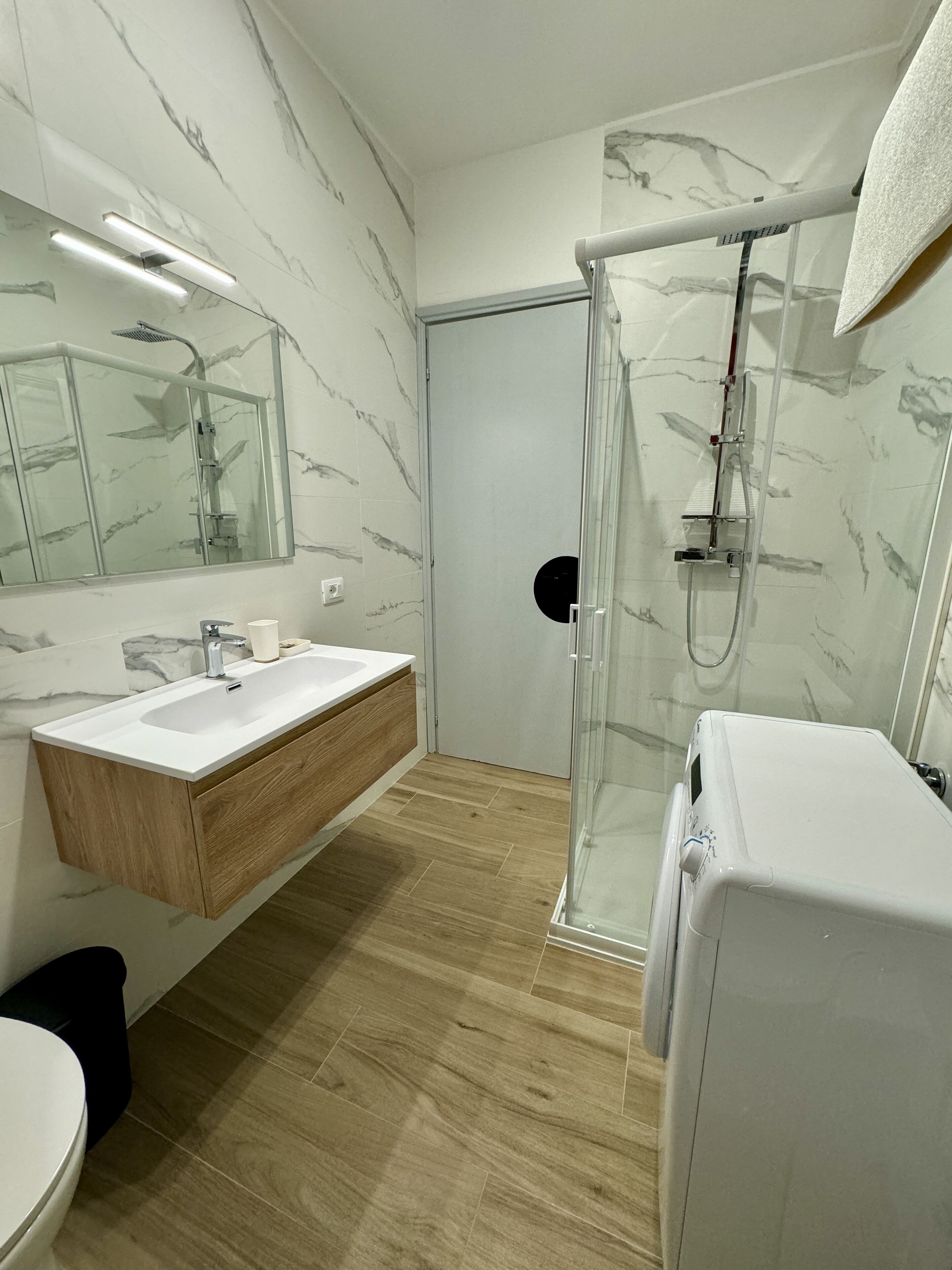 A modern bathroom with a marble backsplash, a wooden vanity, a glass shower enclosure, and a white toilet.