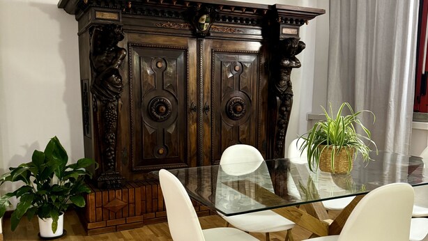 Een eetruimte met een glazen tafel, witte stoelen, een houten kast met houtsnijwerk en een potplant.