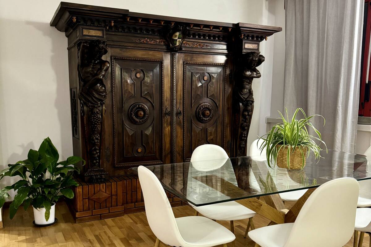 Un comedor con mesa de vidrio, sillas blancas, un aparador de madera con detalles tallados y una planta en maceta.