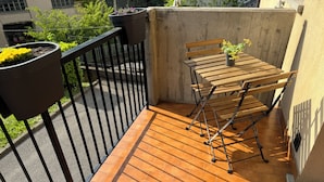 Een klein balkon buiten, met een houten tafel en stoelen, planten in potten en een metalen hekwerk.