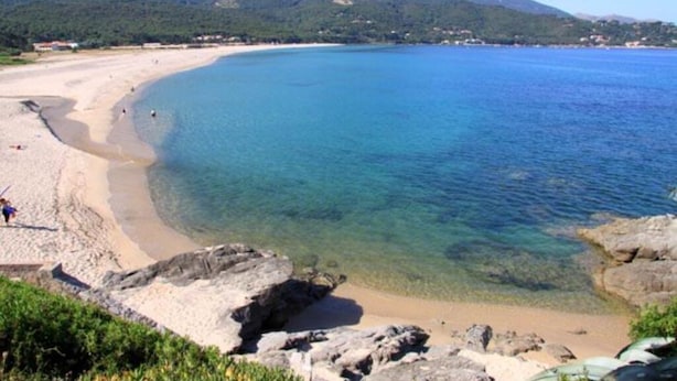 Una playa costera con aguas cristalinas, afloramientos rocosos y montañas al fondo.