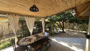 Un espace extérieur aménagé pour les repas, avec une table ronde et des chaises, un luminaire suspendu et un patio couvert doté d’un plafond en bambou.