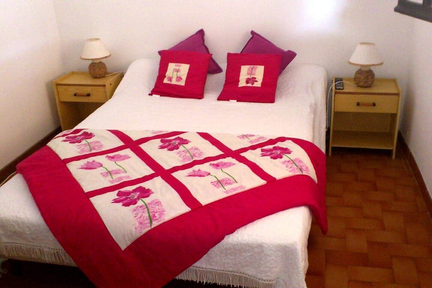 Un lit recouvert d’une courtepointe à motifs floraux, deux oreillers violets et deux tables de chevet avec des lampes.