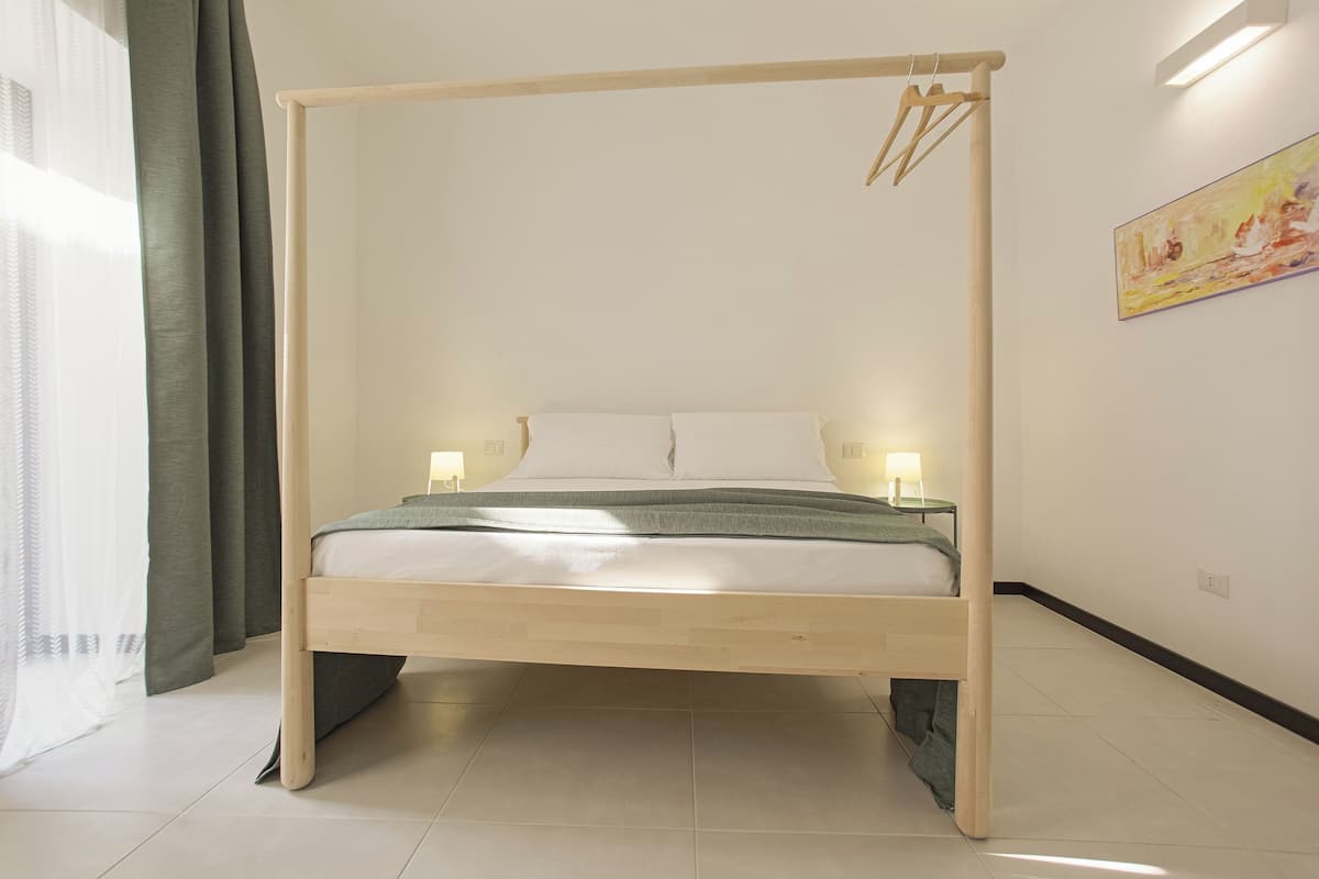 A bed with a wooden frame and a canopy, two bedside lamps, a painting on the wall, and a window with curtains.