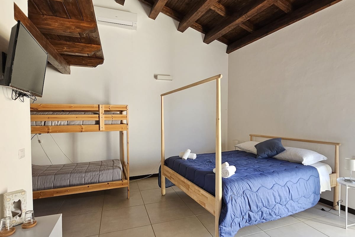 A room with a bunk bed, a single bed, a desk with a chair, and a television on the wall.