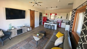 Ein modernes Wohnzimmer mit einer grauen Couch, einem gemusterten Teppich und einem Fernseher. Im Hintergrund ist eine Küche mit roten Geräten und ein Essbereich mit Hockern zu sehen.