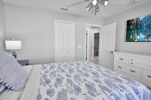 A bedroom with a bed, a nightstand, a lamp, a ceiling fan, a door, a dresser, and a painting. - 4BR Cape | Solar Pool | Bikes Included (Cape Coral)