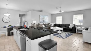 A modern kitchen with a central island, a dining area with chairs, a sofa, and a coffee table. There is a clock on the wall and a painting above the sofa. - 4BR | Solar-Powered Heated Pool | Sleeps 8 (Cape Coral)