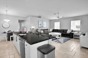 A modern kitchen with a central island, a dining area with chairs, a sofa, and a coffee table. There is a clock on the wall and a painting above the sofa. - 4BR | Solar-Powered Heated Pool | Sleeps 8 (Cape Coral)