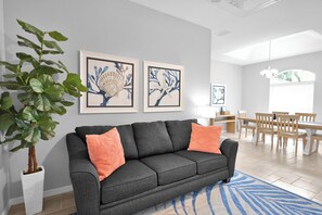 Un salon moderne avec un canapé gris, deux coussins orange, un tapis bleu et blanc, et deux œuvres d’art encadrées accrochées au mur.