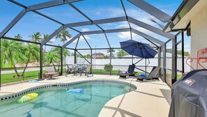 A covered outdoor swimming pool with lounge chairs and an umbrella, surrounded by palm trees. - 4BR | Solar-Powered Heated Pool | Sleeps 8 (Cape Coral)