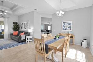 A modern living room with a grey sofa, a dining table with chairs, and a kitchen in the background. - 4BR Cape | Solar Pool | Bikes Included (Cape Coral)