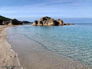 A beach with clear water, a rocky outcrop, and a distant view of the sea.