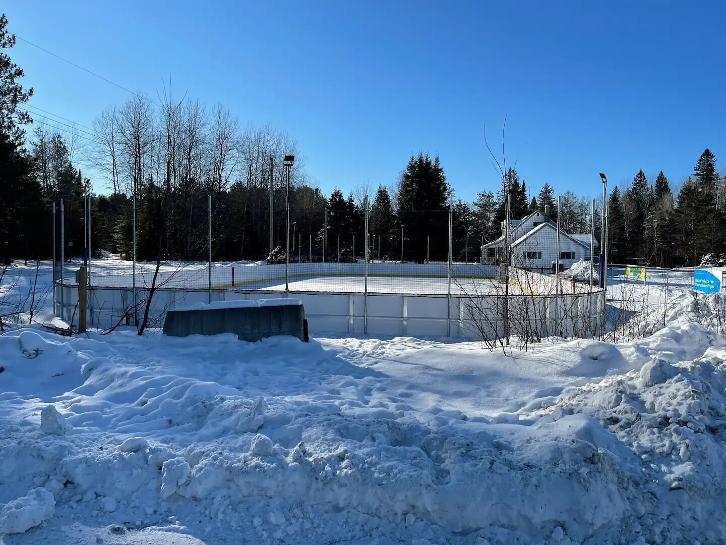 A snow-covered outdoor area with a fenced hockey rink, a building with a sloped roof, and a clear blue sky.