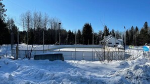 Un espace extérieur enneigé avec une patinoire entourée de barrières, un bâtiment doté d’un toit en pente et un ciel bleu dégagé.