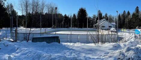 A snow-covered outdoor area with a fenced hockey rink, a building with a sloped roof, and a clear blue sky.