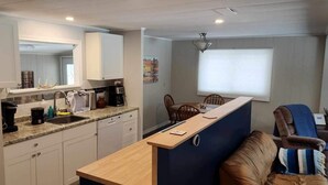 Une cuisine compacte comprenant un évier, un plan de travail et des placards. On y trouve également un coin repas avec une table et des chaises. Une fenêtre avec des stores est visible.