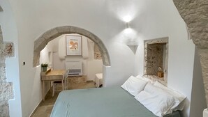 Un dormitorio con un arco de piedra, una cama con cubrecama verde, un escritorio con silla y una mesita con una planta.